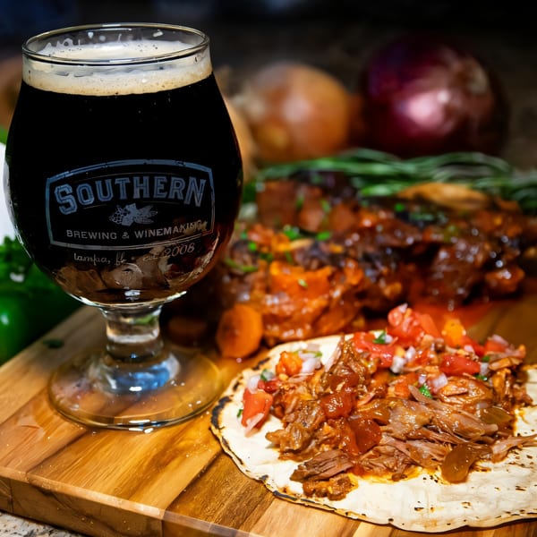 A dark beer in a branded glass next to a Mexican taco with meat and salsa on a wooden board.